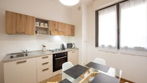 A compact kitchen with a microwave, sink, and wooden cabinets. There is a dining area with a glass table and white chairs. - U Genova (Genoa)