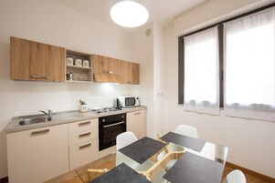 Cocina compacta con microondas, pileta y muebles de madera.  Hay una zona de comedor con una mesa de vidrio y sillas blancas.