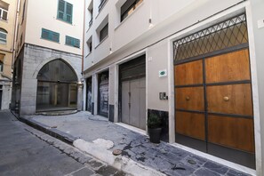 A narrow street with a building featuring a large arched entrance, a garage with a wooden door, and a small potted plant.