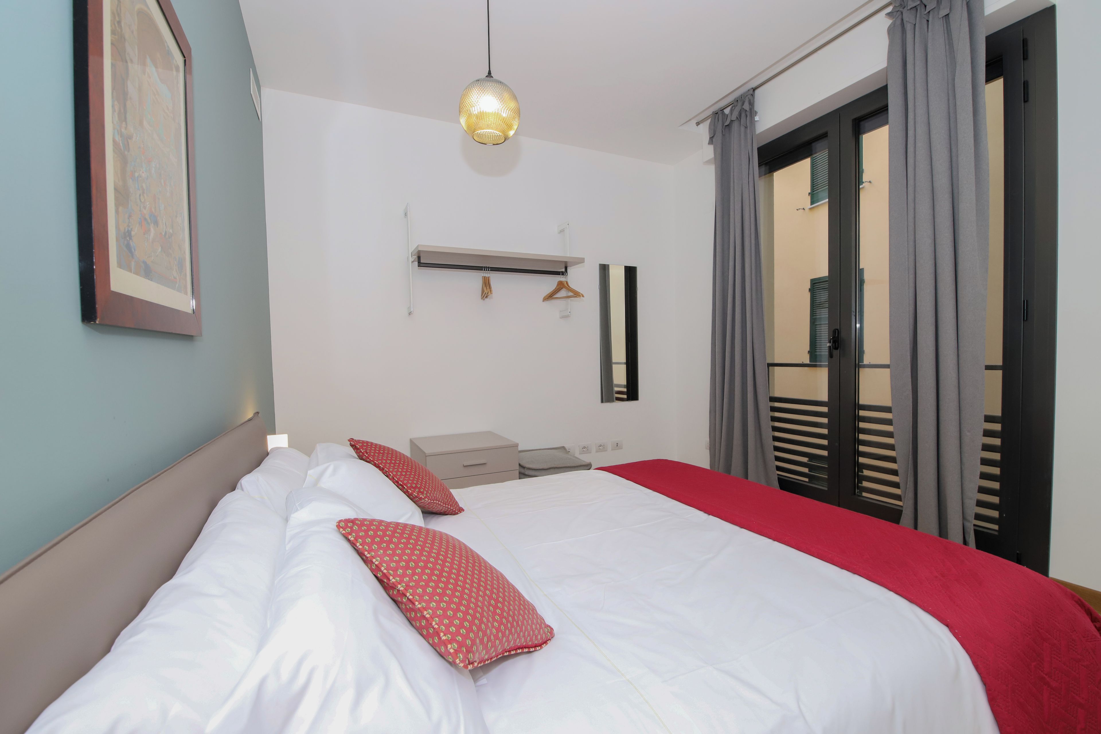 A hotel room with a bed, a bedside table, a hanging light fixture, a mirror, and a view of the outside through a door.