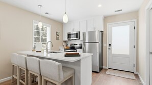 A modern kitchen with white cabinetry, stainless steel appliances, a central island with bar stools, and pendant lighting. - Treehouse at BootLegger Cove (Bluffton)