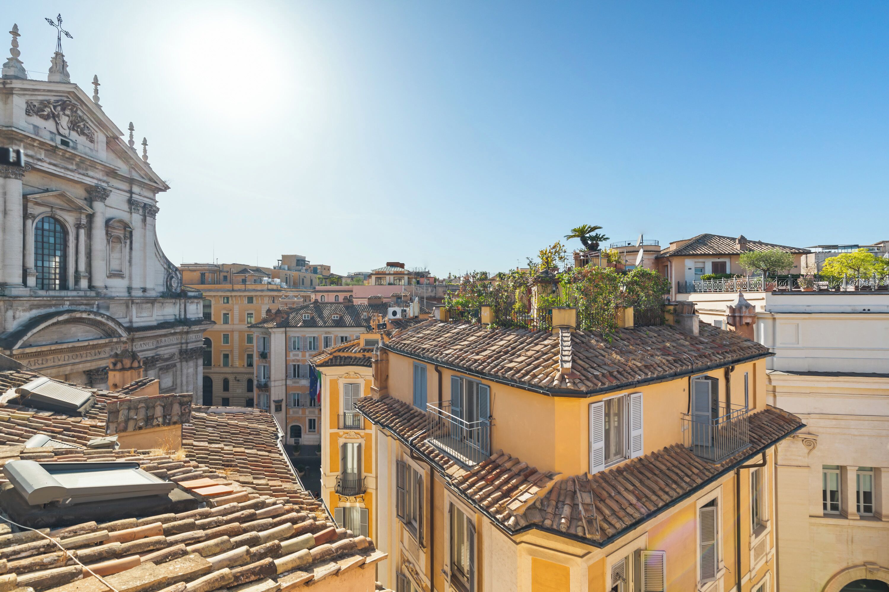 A cityscape with a prominent historical building, terracotta-roofed buildings, and a clear sky.