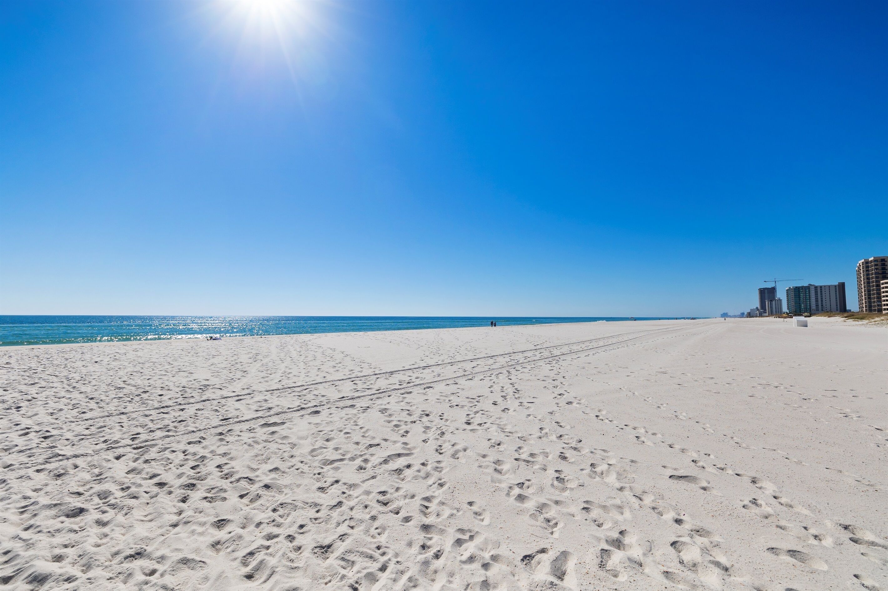 A wide sandy beach with footprints leading to the water, a clear blue sky, and high-rise buildings in the background.