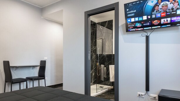 Una habitación de hotel moderna con una cama, un escritorio con dos sillas, un box de ducha con paredes de cristal y un televisor montado que muestra diferentes canales.