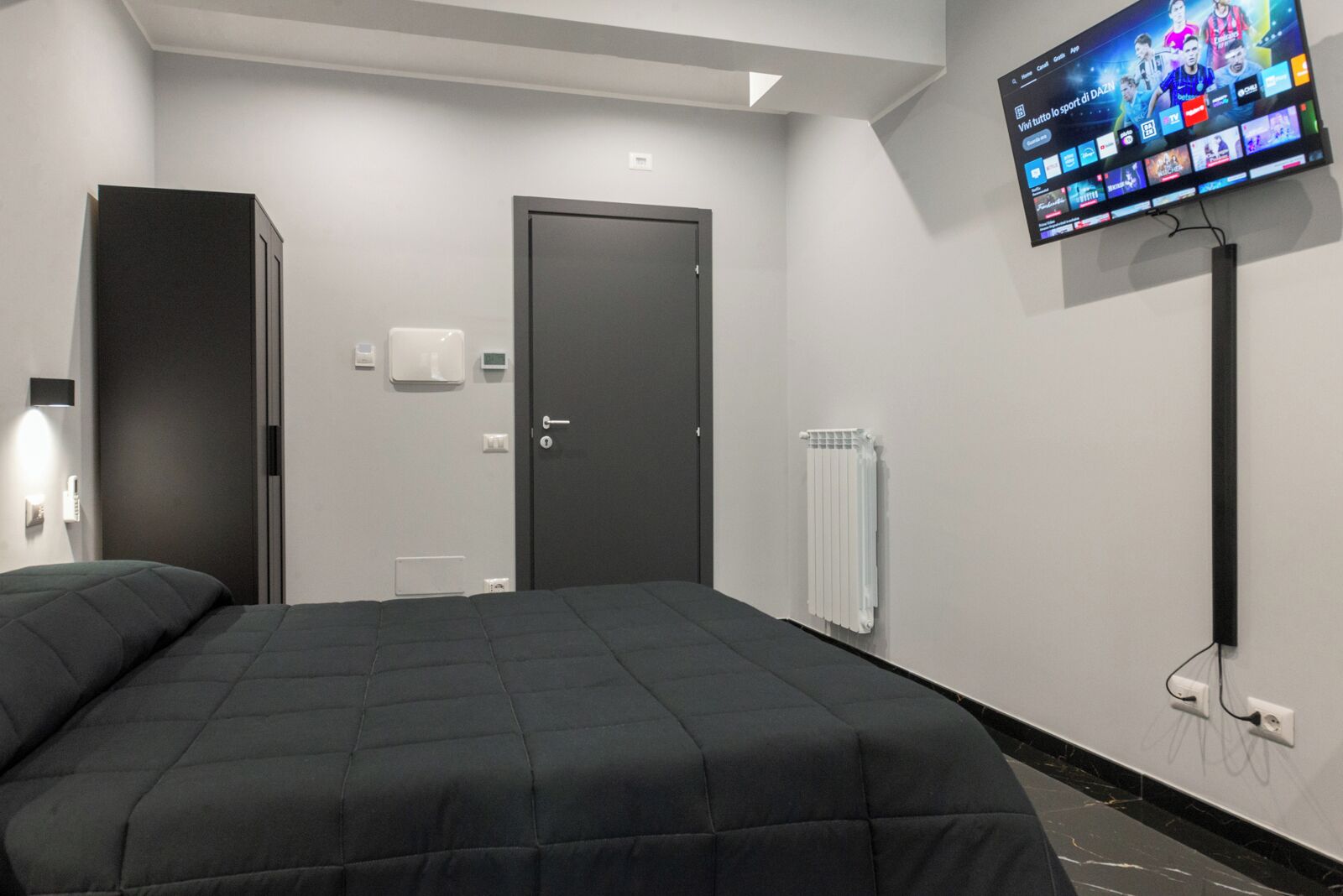 Quarto de hotel moderno com uma cama grande, uma TV de tela plana na parede, uma porta e um radiador.