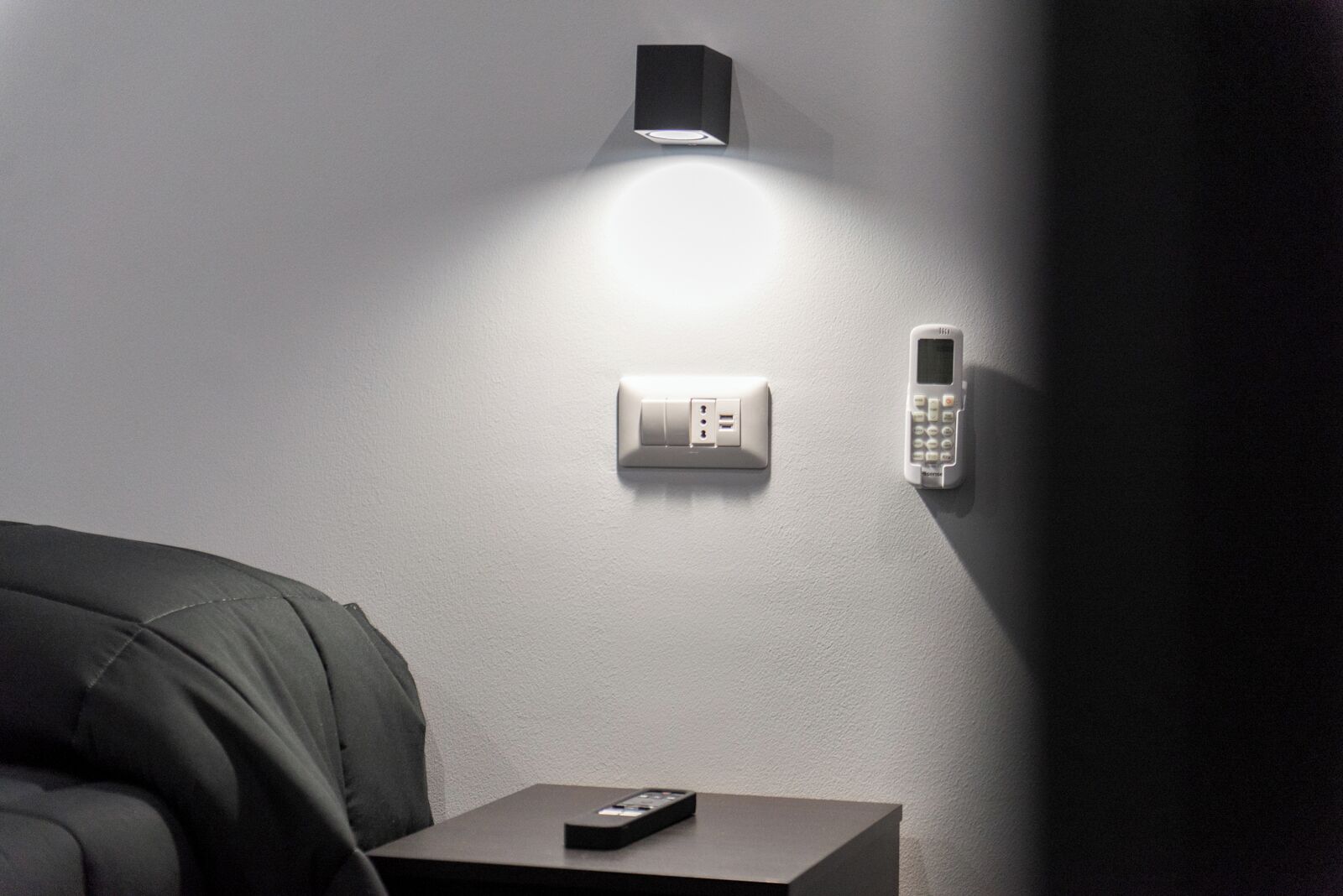 Ein modernes Zimmer mit einem Bett, einem Nachttisch mit Fernbedienung, einem wandmontierten Thermostat und einem an der Wand befestigten Telefon.