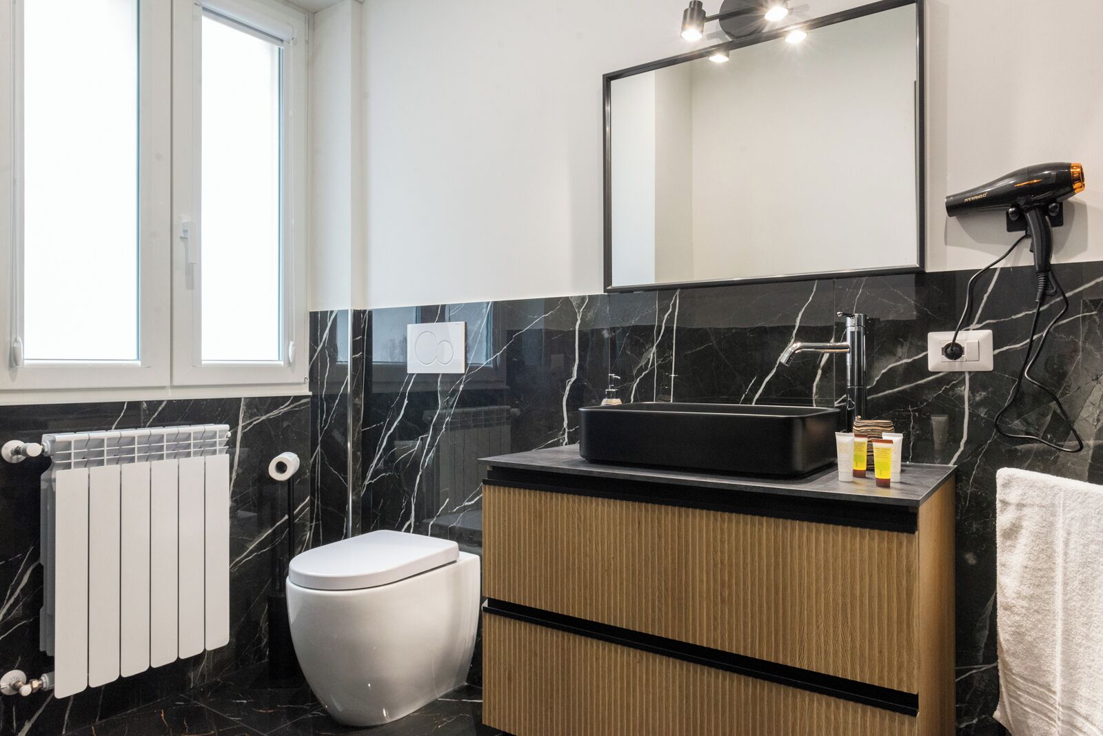 A modern bathroom with a black and white marble backsplash, a white toilet, a wooden vanity with a black sink, and a hairdryer mounted on the wall.