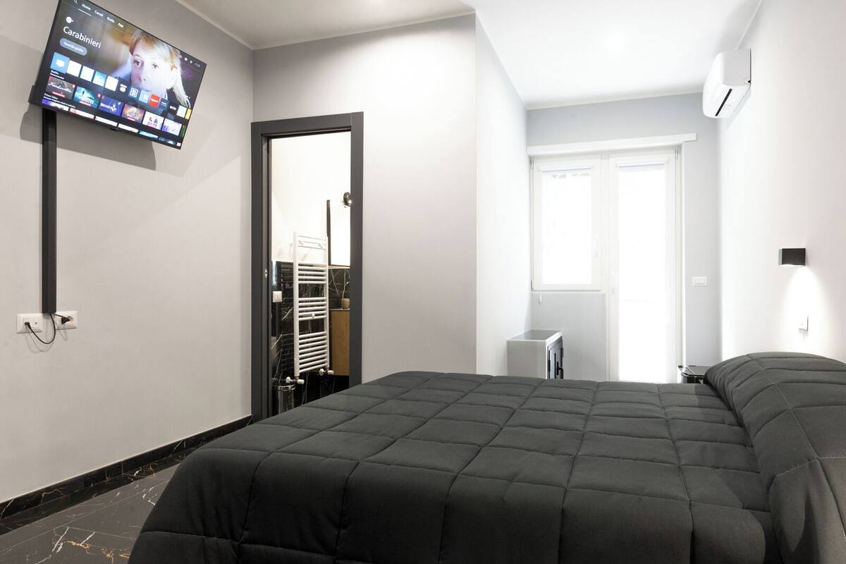Une chambre d’hôtel moderne dotée d’un grand lit, d’un téléviseur à écran plat fixé au mur, d’une petite table de chevet et d’une fenêtre donnant sur l’extérieur.
