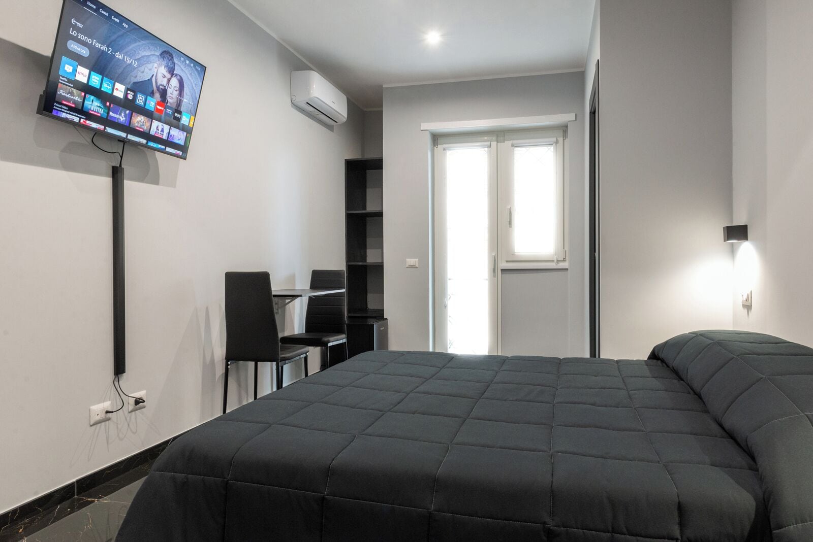 A modern hotel room with a large bed, a desk with chairs, a flat-screen TV mounted on the wall, and an air conditioning unit.