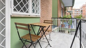 Un balcón con una mesa de madera y dos sillas, barandilla blanca y vista de edificios cercanos.