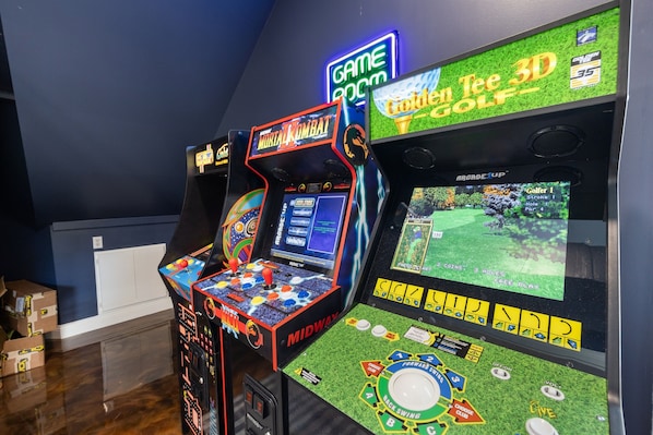 A gaming area with arcade machines, including a 3D golf game and Mortal Kombat, in a room with a blue wall and wooden flooring.