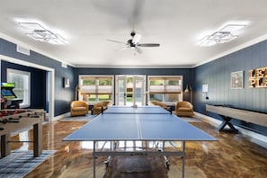 A room with a ping pong table, foosball table, and a pool table. There are two windows with a view of the outdoors.