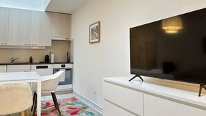 Et kompakt køkken med hvide skabe, et spiseområde med et hvidt bord og stole, og et stort fladskærms-tv monteret på væggen.
