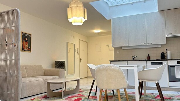 En moderne stue med sofa, lille bord og stol. Der er et køkkenområde med skabe og bordplade. En dekorativ måtte ligger på gulvet.