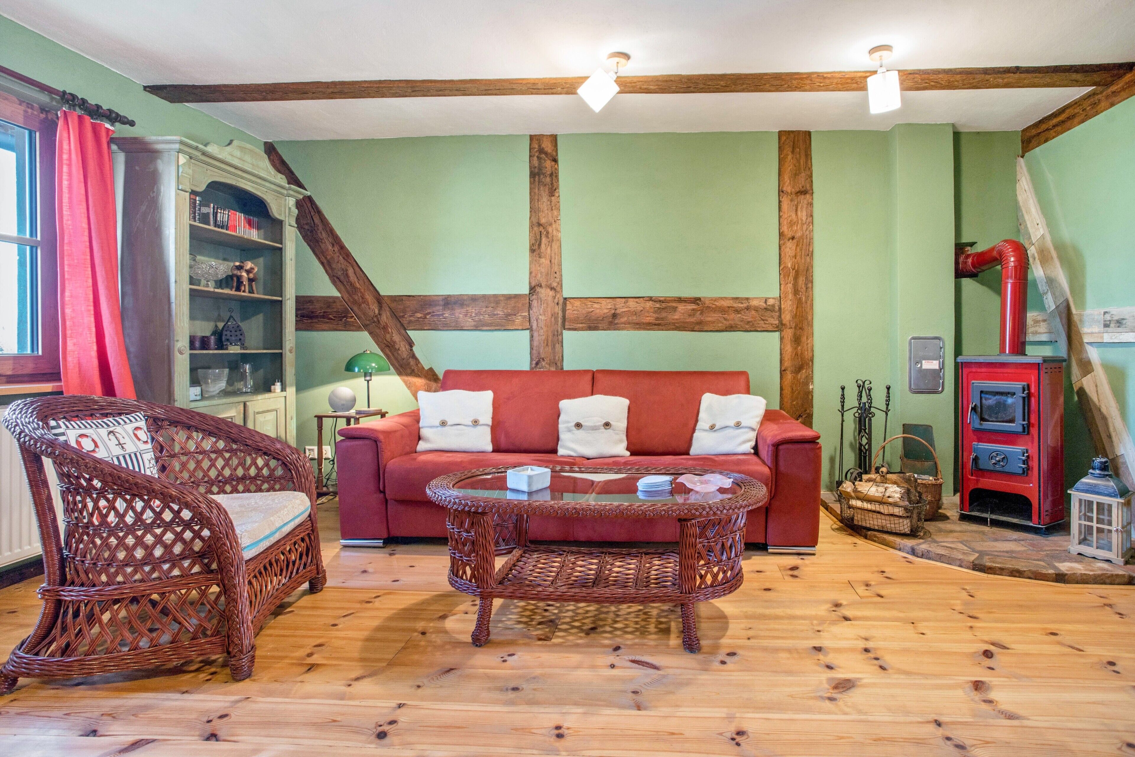 Ein Zimmer mit einem roten Sofa, Korbstühlen, einem Glastisch, Holzboden und einem roten Herd.