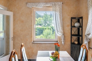 Un coin repas comprenant une table, des chaises, une fenêtre avec des rideaux et une vue sur la verdure à l’extérieur.