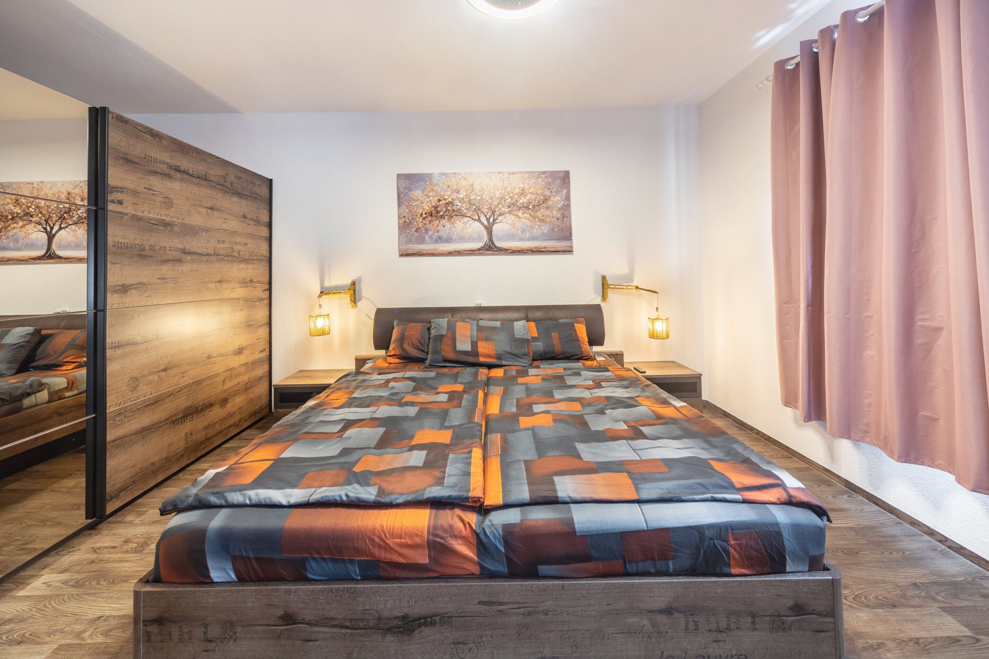 Une chambre moderne avec un grand lit, deux lampes de chevet, une tête de lit en bois, un tableau représentant un arbre et une porte coulissante.