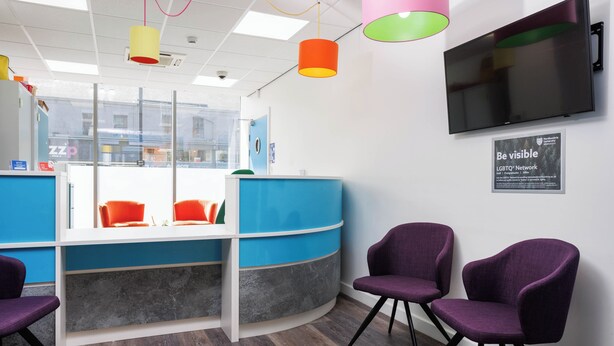 A reception area with a blue counter, purple chairs, a mounted TV, and a 'Be visible' sign.