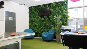 A room with a green wall, a pool table, two blue chairs, a ping pong table, and a TV mounted on the wall.