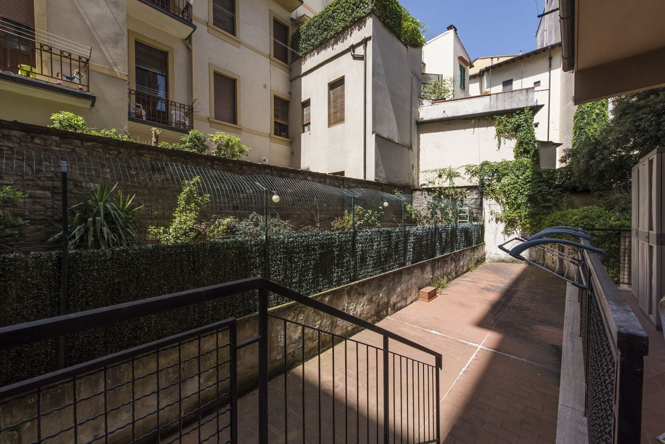 A courtyard with a stone wall, a metal fence, and a staircase leading to an upper level.