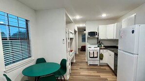 Cocina compacta con armarios blancos, un microondas, una estufa y un refrigerador. Hay una zona de comedor con una mesa y sillas verdes.