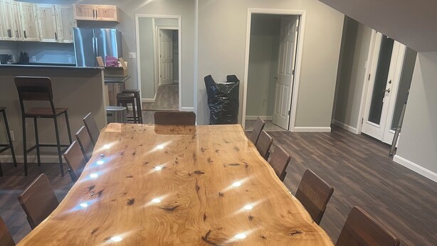 Une grande table à manger en bois, avec des chaises, un coin cuisine équipé de placards et d’appareils électroménagers, et un couloir menant à une porte.