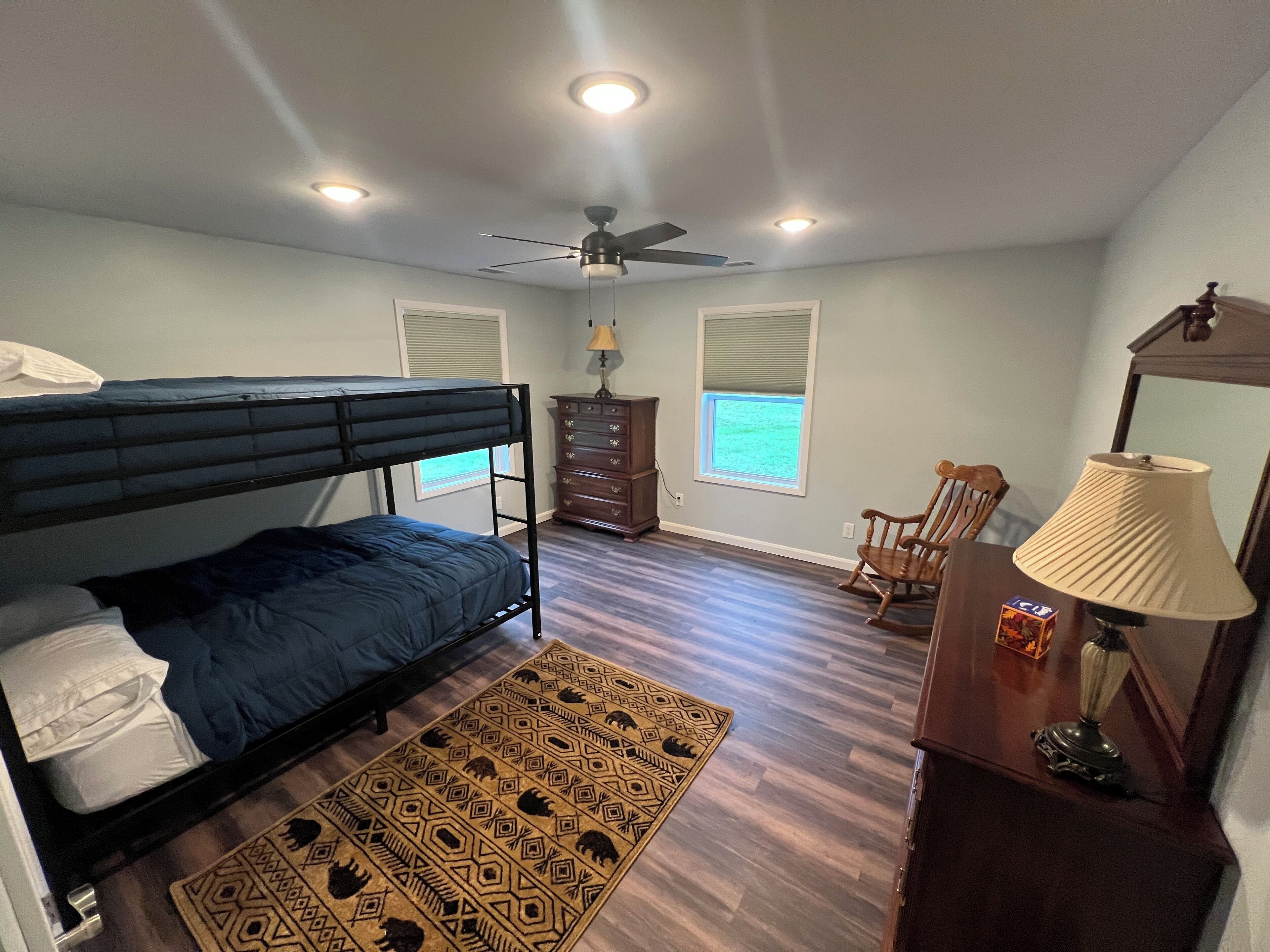 A bedroom with a bunk bed, a desk with a lamp, a chair, a dresser, and a window with blinds.