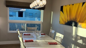 Un comedor con una larga mesa, sillas, una araña y un ventanal grande, además de un cuadro de un girasol en la pared.