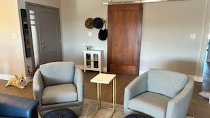 Una sala de estar con dos sillones grises, una mesita blanca y un otoman azul oscuro. Se ve una puerta de madera y un armario blanco con sombreros al fondo.
