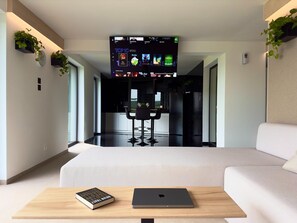 Un salon moderne avec un téléviseur à écran plat affichant une liste de films, un canapé, une table basse avec un livre et un ordinateur portable, et un coin cuisine à l’arrière-plan.