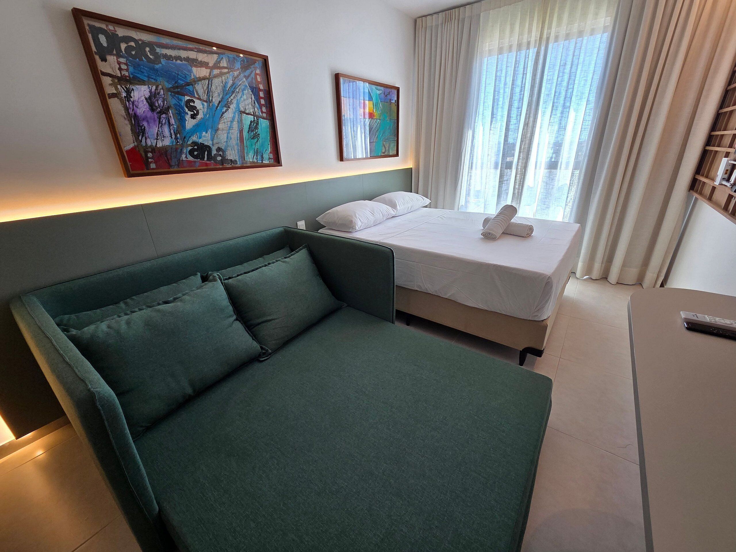 A modern hotel room with a green sofa, a bed with white linens, and two abstract paintings on the wall.