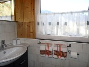 Ein Badezimmer mit Waschbecken, einem Fenster mit durchsichtigen Vorhängen und zwei Handtüchern an einem Haken.