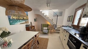 Et moderne køkken med en central ø, et spiseområde med fletstole, en trappe og havudsigt gennem et vindue.