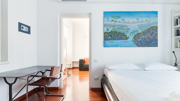 A room with a bed, a desk with a chair, a painting on the wall, and a door leading to another room. - GuestHost - Cozy 50m2 apartment, perfect for 2 people, located on the first floor of a building with lift.The accommodation is located in the Derganino area, about 600 meters from the Lancetti railway station and just over 1 km from the Maciachini  (Milan)
