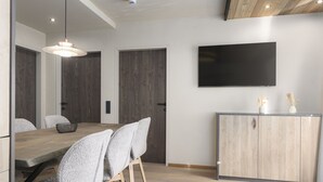 Een moderne eetruimte met een houten tafel, grijs gestoffeerde stoelen, een hanglamp, een flatscreen televisie aan de wand en een houten kastje met een handgreep.