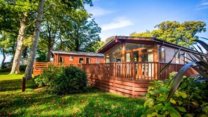 A wooden cabin with a deck, surrounded by lush greenery and trees. - Coed Helen Holiday Park (Caernarfon)