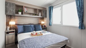 A neatly arranged bedroom with a bed, bedside tables, a nightstand, a window with curtains, and built-in shelves with books and decorative items. - Coed Helen Holiday Park (Caernarfon)