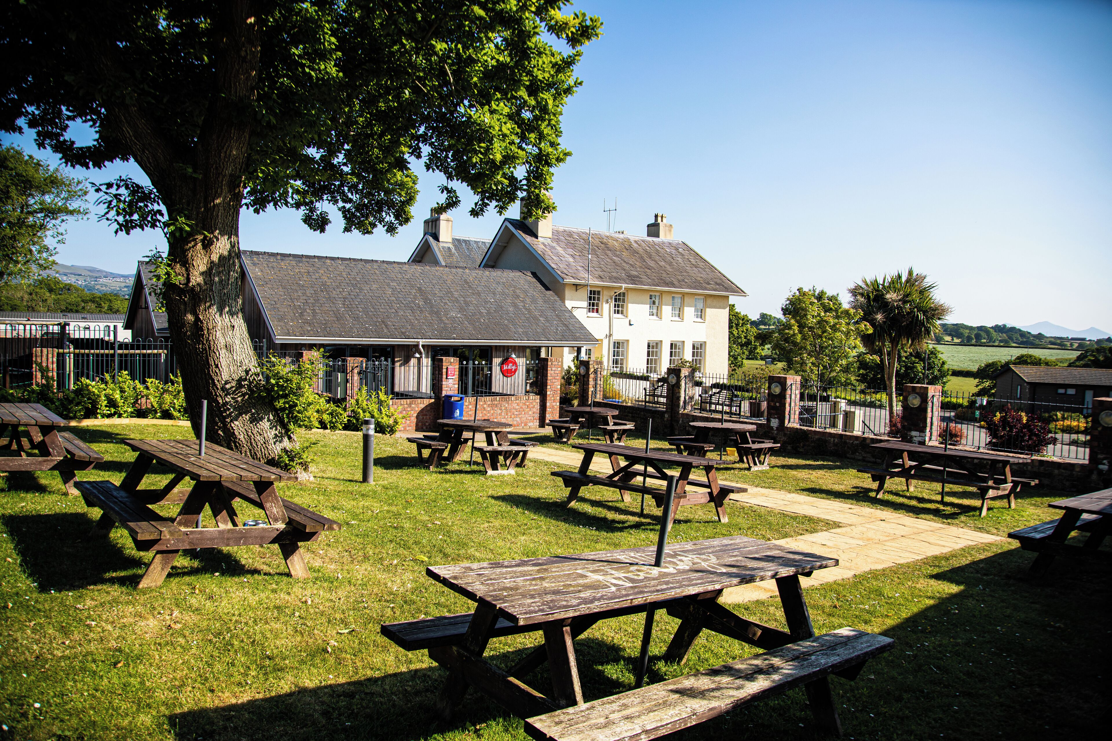 Een picknickplaats met meerdere houten tafels en banken, omgeven door een grasveld en een gebouw met een schuin dak.
