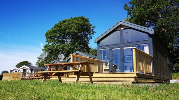 A row of wooden cabins with large windows, a picnic table, and a grassy area. - Coed Helen Holiday Park (Caernarfon)