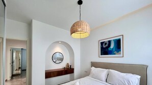 Una habitación de hotel moderna con una cama grande, un espejo redondo, una cómoda de madera y una lámpara colgante.