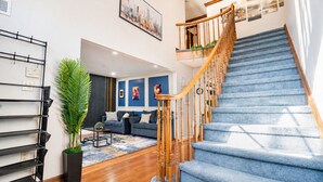 A staircase with wooden railings, a blue carpeted floor, a dark blue sofa set, and a cityscape painting on the wall. - Mega Mansion! 6BR-3.5BA | NYC | MetLife | Parking (Bayonne)