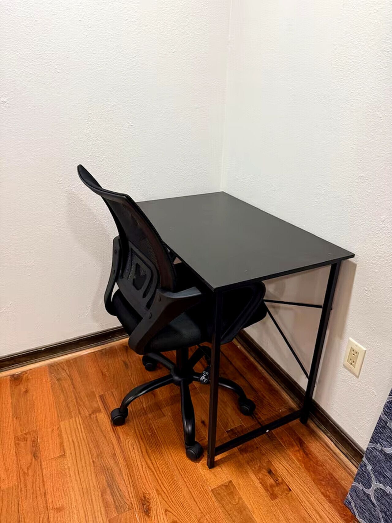 Een hoekbureau met een zwart bureau en stoel, houten vloer en een witte muur.