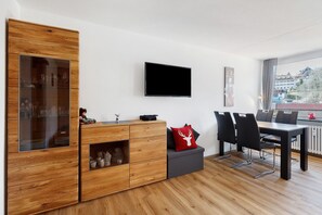 Moderni olohuone, jossa on puinen kaappi, litteänäyttöinen televisio, ruokapöytä tuoleineen ja ikkuna, josta on näkymä rakennuksiin.