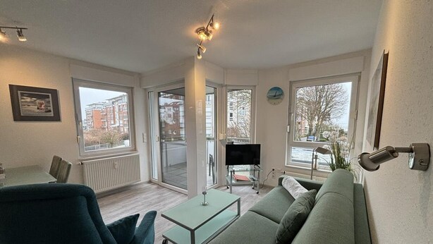 Ein modernes Wohnzimmer mit einer Couch, einem Couchtisch und einem Fernseher. Ein Balkon bietet Blick auf Gebäude, und eine Tür führt nach draußen.