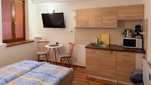 A compact room with a bed, a small dining table with chairs, a kitchenette with a microwave, and a TV mounted on the wall.