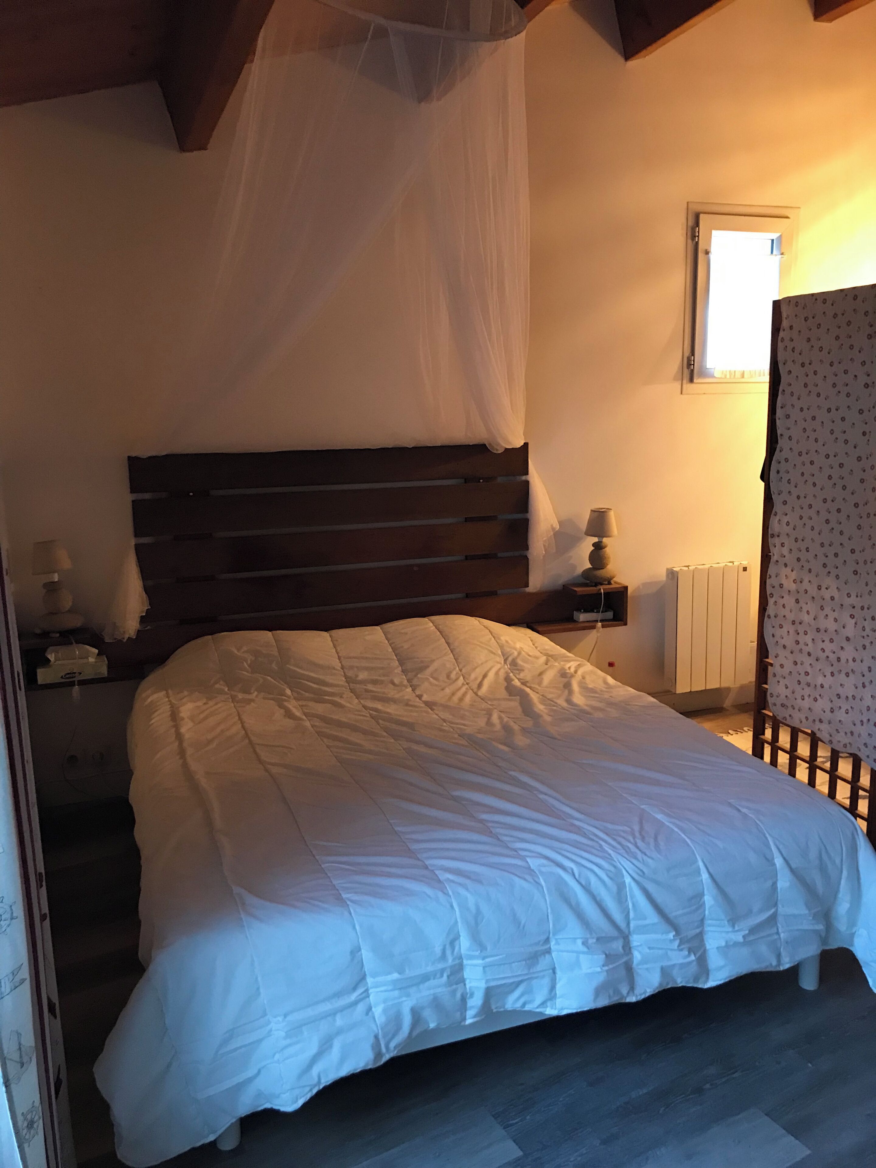 A bedroom with a large bed, a wooden headboard, a bedside table with a lamp, a window with curtains, and a radiator.
