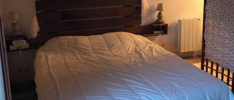 A bedroom with a large bed, a wooden headboard, a bedside table with a lamp, a window with curtains, and a radiator.