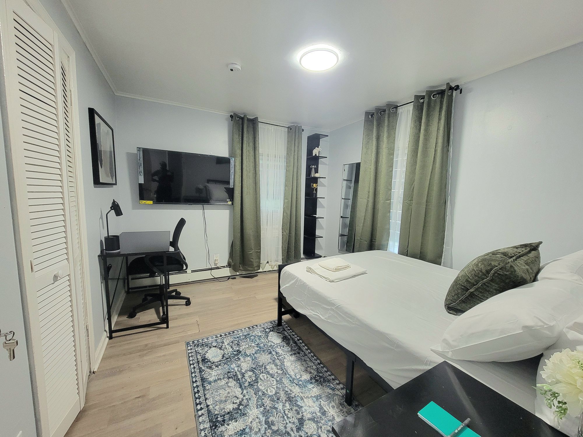 Een slaapkamer met een bed, een bureau, een stoel, een televisie en een raam met gordijnen.