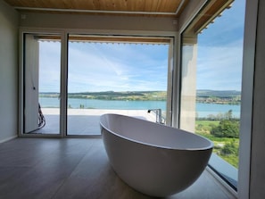 Une salle de bain moderne avec une baignoire îlot, de grandes portes coulissantes en verre et une vue sur un lac et des collines.