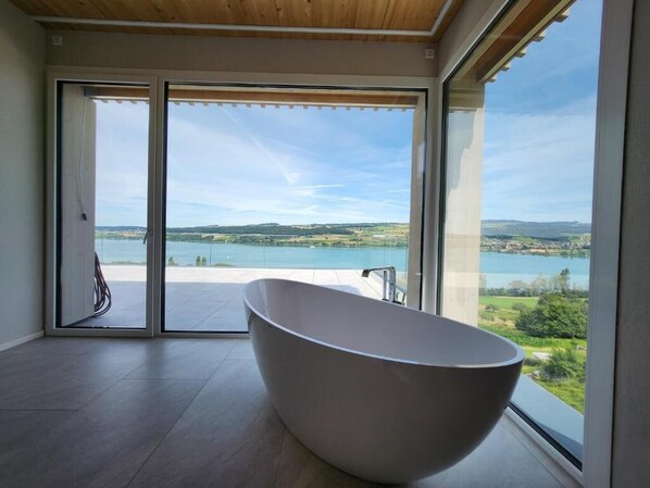 A modern bathroom with a freestanding bathtub, large sliding glass doors, and a view of a lake and hills. - Lakeview Guest Rooms Rooftop Terrace (Ebikon)